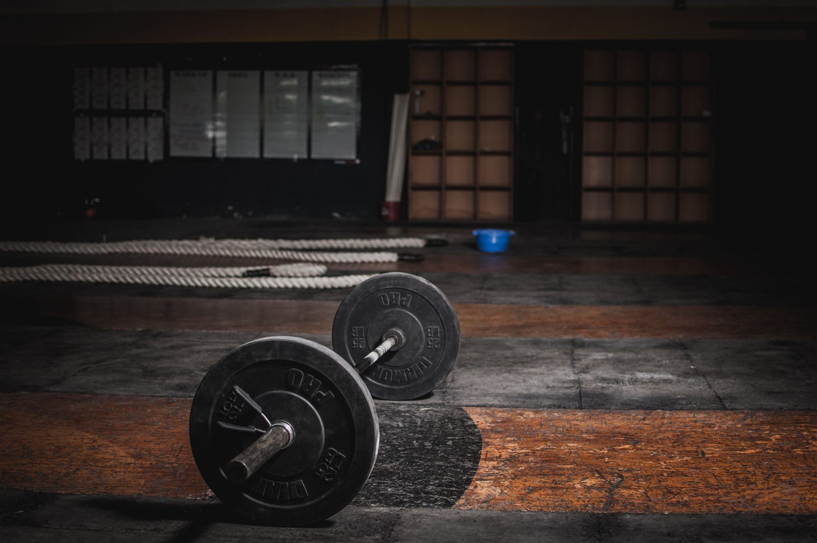 A loaded barbell on a gym floor in low light, with the room behind it in shadow